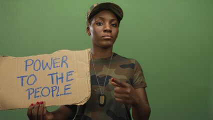 Woman soldier wearing camouflage uniform and cap points finger to cardboard sign in studio;...