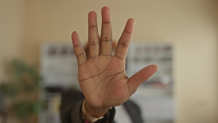 Obraz premium Young african american woman raising open palm to camera in a modern office setting, conveying a clear stop gesture; assertiveness.