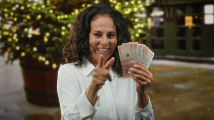 Middle-aged woman holding colombian pesos on a city street smiling under festive lights outdoors...