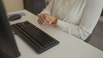 Woman rubs wrist on white desk beside black computer keyboard and monitor in office building; discomfort.