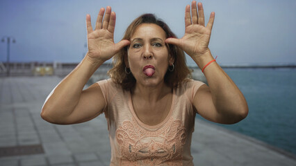 Middle aged woman in peach blouse raises palms beside head on seaside concrete promenade; playful...