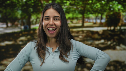 Obraz premium Young hispanic woman smiling in a park surrounded by green trees enjoying the sunny outdoors with a joyful expression and casual attire.