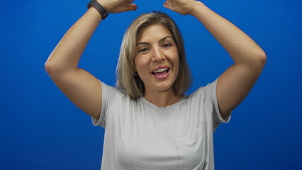 Woman wearing white t shirt lifts bare arms over head with a bright smile in blue studio setting;...