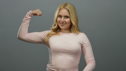 Woman flexing muscles confidently against a smooth grey background, wearing a casual pink shirt,...