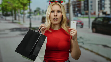 Young blonde woman in red shirt holding shopping bags and card on city street, portraying surprise outdoors.