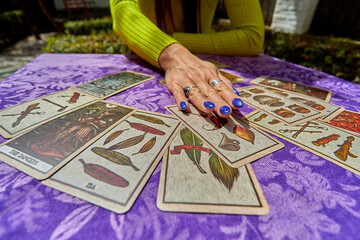 A woman with bright blue nails lays tarot cards on a purple cloth, revealing symbols of feathers,...