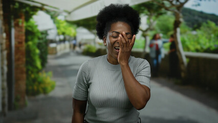 Fototapeta premium Woman laughing with hand on face outdoors on a city street with greenery in the background, capturing a candid moment of joy.