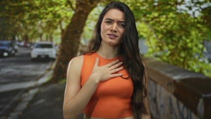 Young hispanic woman in orange crop top with long hair touches her chest against graffiti wall on city street outdoors; indignation.