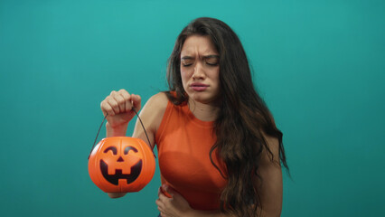 Young hispanic woman holding pumpkin bucket and clutching stomach in studio with teal background;...