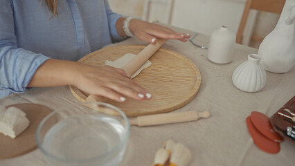 Woman rolls clay slab on a round wooden board with rolling pin in studio with natural daylight; creative concentration.