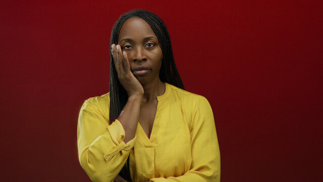 Woman wearing yellow blouse standing against vivid red background displaying thoughtful expression with hand on face, conveying emotions and solitude.