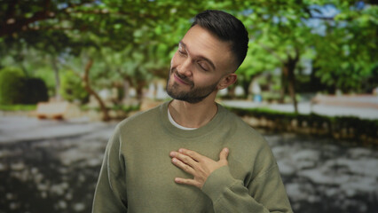 Fototapeta premium Young hispanic man standing outdoors in a green park, smiling warmly, hand on chest, surrounded by lush trees, exuding calm and peaceful vibes, dressed casually.