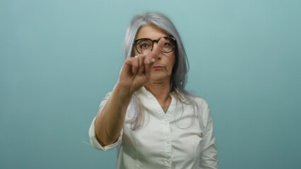 Senior woman with grey hair gesturing no with her fingers against a blue background, asserting a...