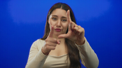 Woman making a loser sign gesture with her hand framing her face in a blue studio setting with...