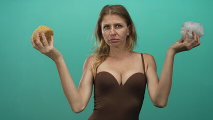 Woman in brown swimsuit holds a yellow sponge and white loofah in teal studio, arms raised and...