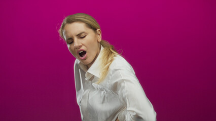 Woman grimacing in pain with hand on back against a vibrant pink background, highlighting discomfort and anguish in an isolated setting.