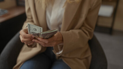 Woman counting stack of us dollar bills in modern office setting, hands focused, wearing beige blazer, highlighting financial themes in a professional indoor environment.