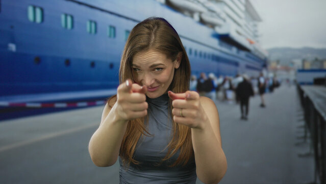 Woman with bare arms points finger at camera on busy port street beside a large white cruise ship under clear sky; playfulness.