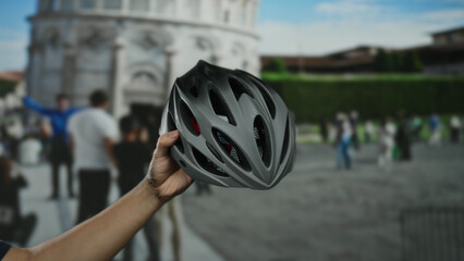 Man holding a bicycle helmet near pisa tower in italy, with a bustling city street and tourists in the background.