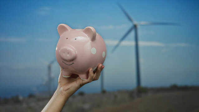 Caucasian man holds a pink piggy bank against a backdrop of windmills in an outdoor field, symbolizing savings and renewable energy, under a clear blue sky. - Powered by Adobe