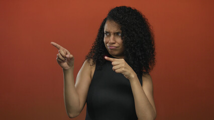 Obraz premium Young hispanic woman with curly hair pointing with both hands against a vibrant red wall showcasing an expressive gesture in a modern studio environment.