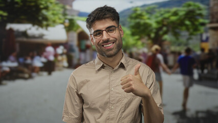 Fototapeta premium Hispanic man wearing glasses and light shirt smiles broadly and gives thumbs up on urban street; approval.