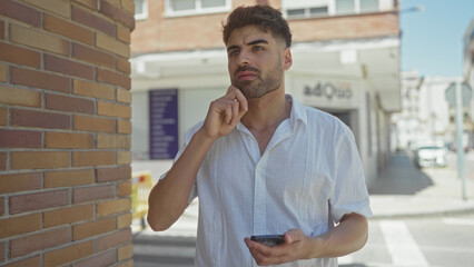 Man taps smartphone with finger on sunlit city street beside brick wall near crosswalk;...
