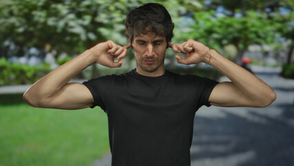 Young man in black t shirt covers ears with fingers under green park canopy beside a paved pathway;...