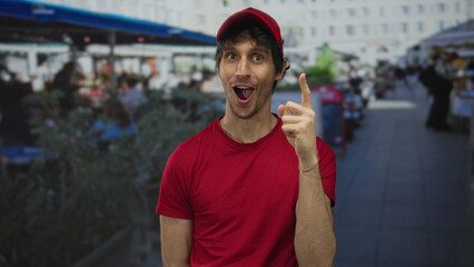 Man in red cap and red shirt points finger to sky on crowded restaurant terrace beside urban street; excitement.
