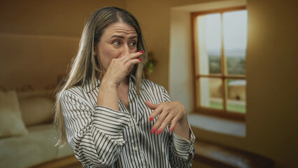 Middle aged blonde woman pinches nose beside window in spacious yellow building lobby wearing...