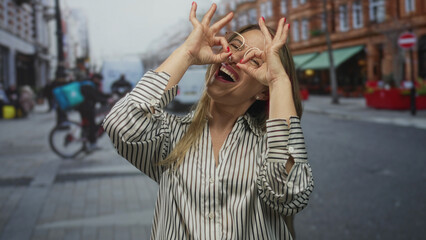 Blonde woman wearing glasses makes ok sign with fingers near eyes on downtown busy city street; playfulness.