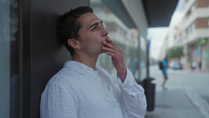 Young man in white shirt stands thoughtfully against a wall on a city street with blurred urban background on a quiet day.