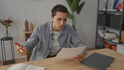 Young man analyzing document at home, sitting in modern living room with laptop and books, conveying focus and concentration, surrounded by stylish decor and personal items.