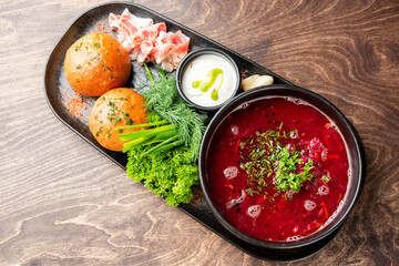 Traditional Eastern European borscht served with sour cream, salo, bread rolls, garlic, and herbs on a rustic wooden table. Top view, vibrant colors, authentic cuisine.
