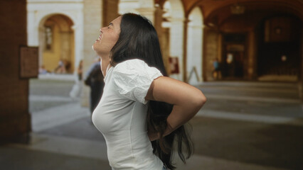 Woman holding lower back in sunlit historic old town stone building; backache discomfort strain tension.