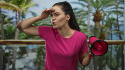 Woman shields forehead with hand and holds pink megaphone while looking into distance in forest setting; determination resolve perseverance courage.