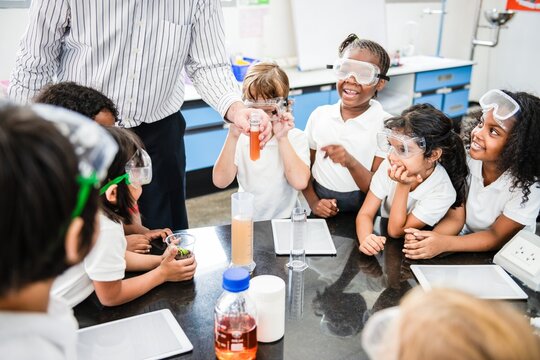 Diverse group of children in a science class, wearing safety goggles, excitedly watching a teacher demonstrate an experiment with colorful liquids in a lab. Diverse young students in science class.
