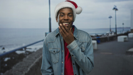 Young man wearing santa hat and denim jacket touches chin and smiles on seaside street under lamp posts; holiday joy.