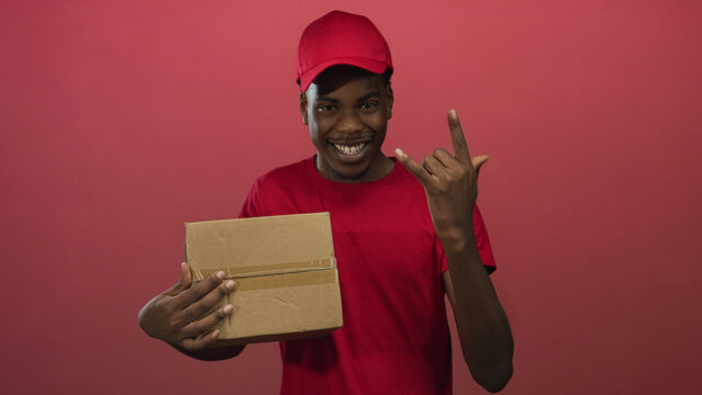 Man wearing red cap and shirt holding cardboard box smiles showing teeth while making rock horns gesture in red studio; enthusiasm.
