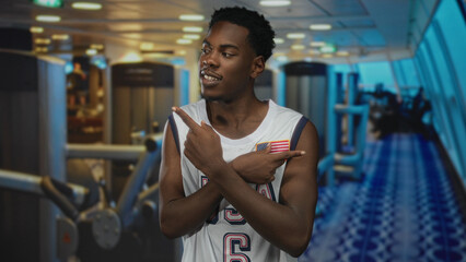 Young african american man in usa basketball jersey with american flag patch pointing fingers in a gym building; pride strength.