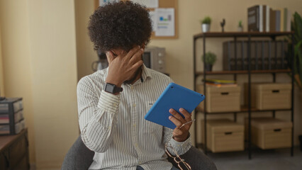 Man in indoor office rubs eyes tired from screen time holding tablet with shelves and files in background.