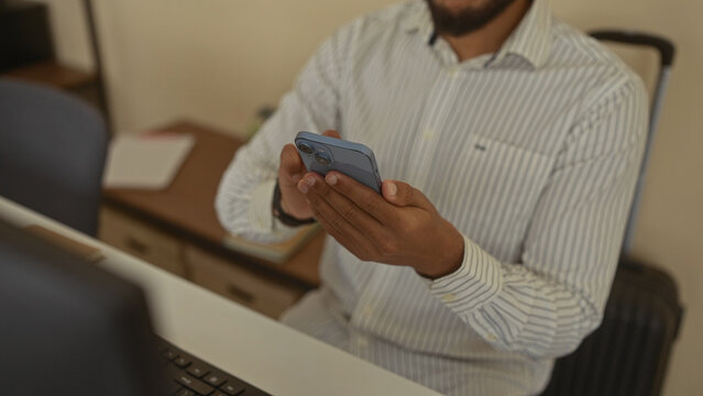 Man using smartphone in office wearing striped shirt checking smartwatch with focused expression indoors desktop background technology scene.