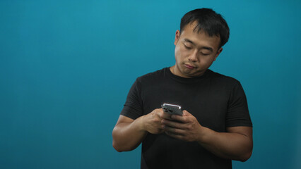 Young chinese man smiling while holding smartphone with both bare hands at chest level in turquoise...