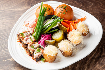 Assorted gourmet appetizers on a triangular plate: baked potatoes with cheese, bread rolls, cured meat, pickled vegetables, dipping sauce, and fresh herbs on rustic wood.