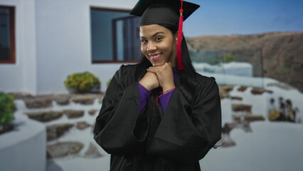Woman in graduation cap and gown with hands under chin gesture at building entrance outdoors; pride...