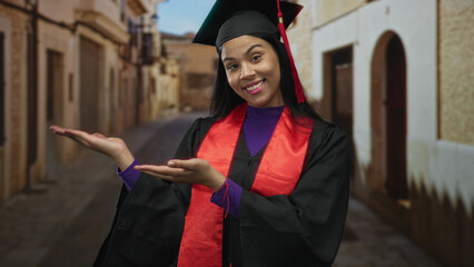 Woman wearing mortarboard presenting open palms on a cobbled old town street; achievement success...