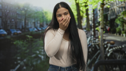 Woman covering mouth with hand while laughing on street by canal water and parked bicycle near building facade; joy.