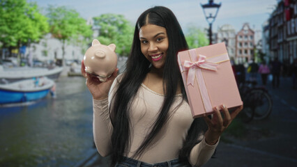Woman holding pink piggybank and pastel giftbox on busy city street beside canal; financial planning optimism.