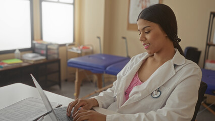 Young latin doctor woman typing on laptop with stethoscope and medical chart at white desk in...