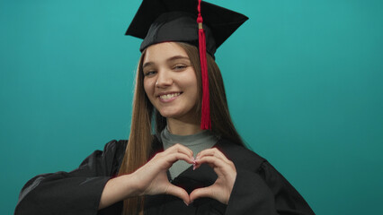 Girl wearing graduation cap makes heart gesture in studio with turquoise background; pride success joy.